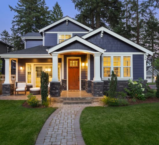 facade of home with manicured lawn, landscaping, and backdrop of trees and dark blue sky. Glowing interior lights create a welcoming mood.